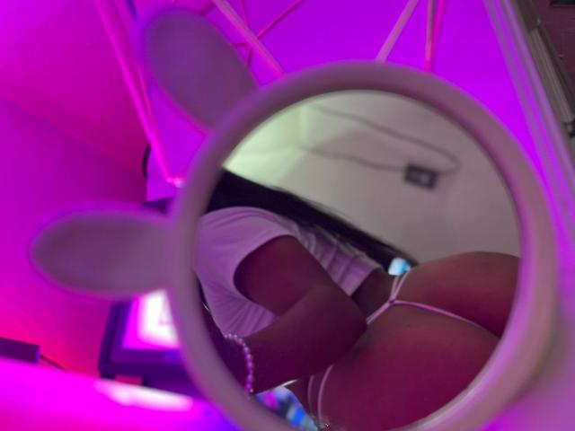 Amberv's ass, reflected in a mirror, showing white underwear and a pink string, with purple and pink neon lights. Her legs are visible, wearing a white bracelet.