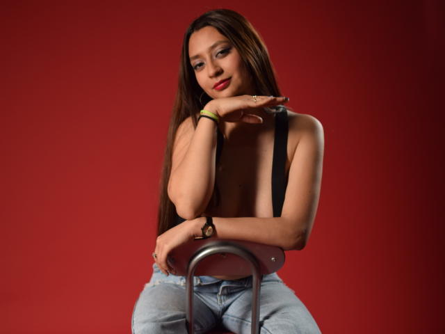 VioletaBlair, com cabelos castanhos longos, sem blusa, usando suspensórios pretos, sentada em uma cadeira, vestindo jeans, batom vermelho, mão direita tocando o queixo, fundo vermelho, sexy, fofa, confiante..