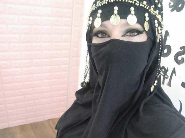 Evelynsalax con burka negra, tocado dorado en forma de moneda y máscara negra que deja ver sus ojos y delineador. Sus glúteos están cubiertos, y su rostro es parcialmente visible..