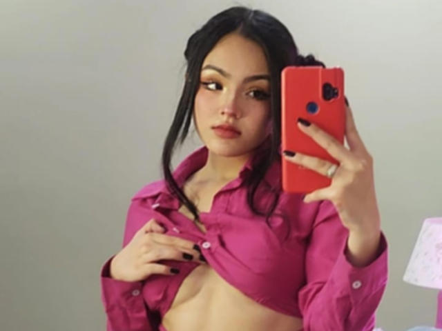 HaleiBeckham macht ein Selfie im Spiegel, trägt ein unknöpftes rosa Hemd, das ihre Brüste zeigt, hat schwarzen Nagellack, hält ein rotes Telefon und schaut in die Kamera..