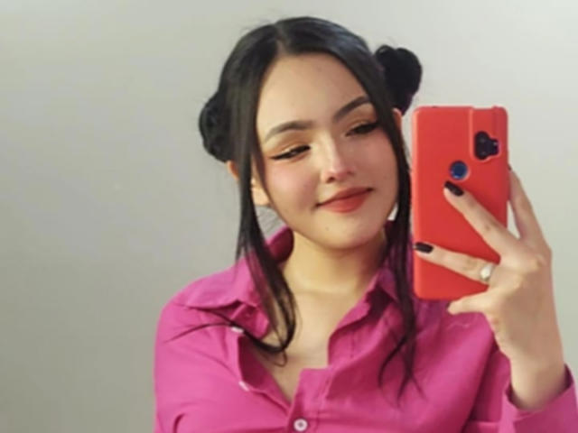 HaleiBeckham con cabello negro en dos moños, camisa rosa, sonriendo, sosteniendo un teléfono rojo con pegatinas de corazón, tomando una selfie, mostrando su esmalte de uñas negro y su cara linda..