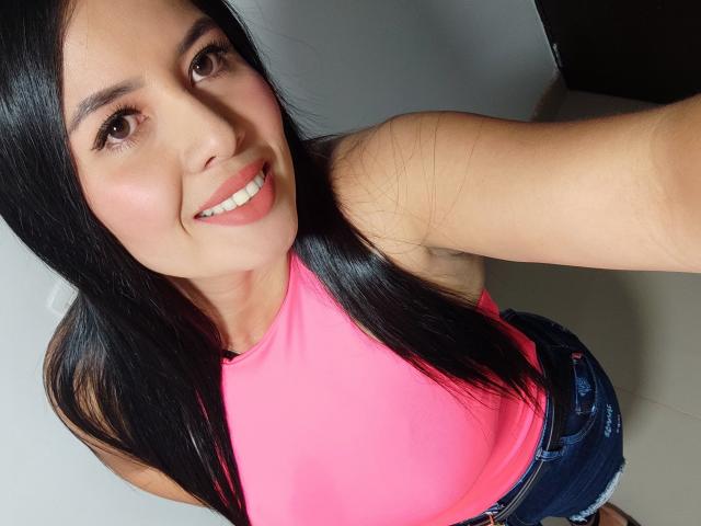 AshleyKatelyn with long black hair, pink top, and denim shorts, smiling at the camera, showing her tits slightly in the bright pink top, and wearing a pink lipstick.