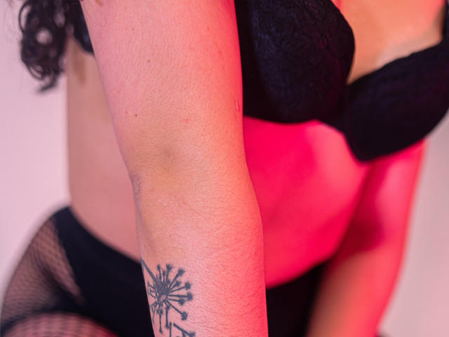 LourrieAnn's arm met een paardenbloem tatoeage, een zwarte beha, een roze top en zwarte onderbroek, waarbij haar blote huid en ronde figuur zichtbaar zijn. Haar borsten zijn gedeeltelijk zichtbaar, ze draagt een netbroek..