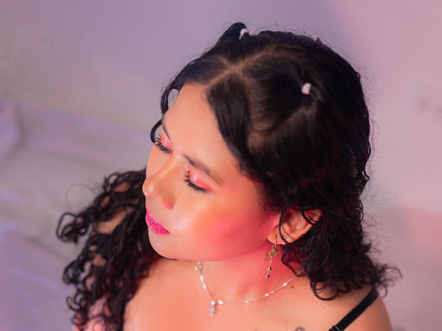 LourrieAnn, com cabelos pretos encaracolados, batom rosa e delineador, olhos fechados, vestindo alças pretas, brincos pequenos e um colar, tem iluminação rosa e roxa no rosto..