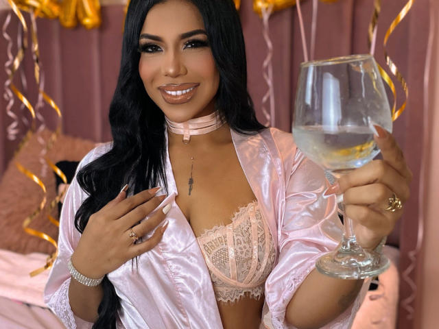 Bella Ivanov mit langem, schwarzem Haar, lächelnd, trägt einen pinken Spitzen-BH und einen glänzenden, pinken Bademantel, hält ein Glas Wasser, trägt eine pinke Halskette und einen goldenen Ring. Brüste sind sichtbar..