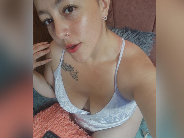 Une femme avec des taches de rousseur, un anneau de nez et des boucles d'oreilles, portant un haut blanc en velours à décolleté plongeant, laissant entrevoir ses seins, avec un tatouage sur l'épaule gauche, assise sur des coussins bleus et orange duveteux..