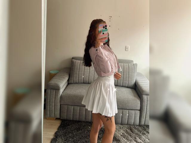 AnyaJay with a pink long-sleeve shirt and white skirt, standing sideways, holding a phone with a green case, covering her tits, brown hair, tattoo on her right thigh.