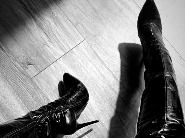 ArleenHunter's shiny black thigh-high boots with high heels and pointed toes, casting shadows on a wooden floor in a black-and-white photo. Boots are zipped up.