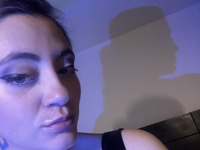 ArleenHunter's face with dark eyebrows, winged eyeliner, and shiny lips, casting a shadow on a wall. She's wearing a black sleeveless top, her eyes are half-closed.