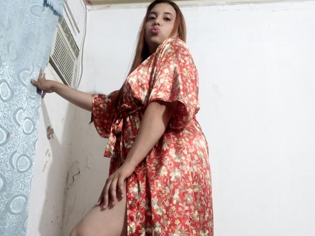 SamiLove mit langen, braunen Haaren, trägt ein rotes Kleid mit Blumenmuster, steht, legt eine Hand an die Wand, blickt nach unten, zeigt ein Bein und macht mit ihren rosafarbenen Lippen eine Schmollmund..