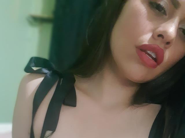 BellaSmithX69 tem batom vermelho, fitas pretas nos seios, e lábios vermelhos ligeiramente entreabertos, com cabelo escuro e um fundo esverdeado. O rosto dela está inclinado para a direita..