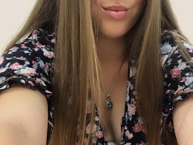 Una mujer con cabello castaño largo, un vestido floral y labios rosados, que lleva un collar con un dije negro, con los labios ligeramente entreabiertos y escote visible..