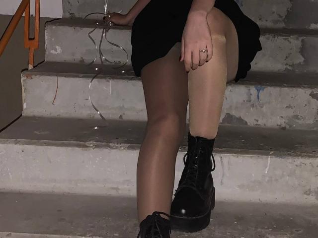 Les jambes de Tserera sont posées sur des marches en béton, elle porte des bottes de combat noires, des collants fins couleur chair et une jupe noire. Sa main droite porte une bague, et des fils sont posés sur les marches..