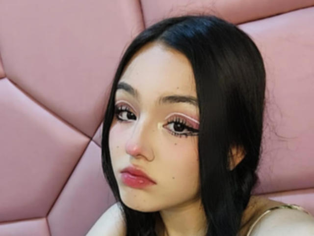 HaleiBeckham hat langes, schwarzes Haar, rosigen Lidschatten und rosafarbenen Lippenstift. Sie schaut leicht schmollend nach unten, trägt ein goldenes Oberteil und hat ein kleines Nasenpiercing..