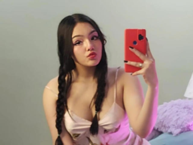 HaleiBeckham with braided dark hair, pouty lips, pink lipstick, wearing a pink dress, holding a red phone, showing her tits, in a mirror selfie.