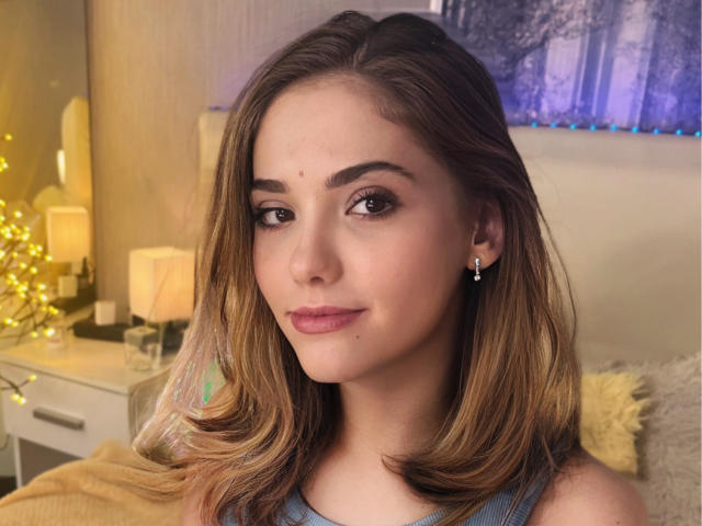 DellaRowe has light brown hair, brown eyes, and a small beauty mark above her right eyebrow. She's wearing a blue top, earrings, and has a slight smile.