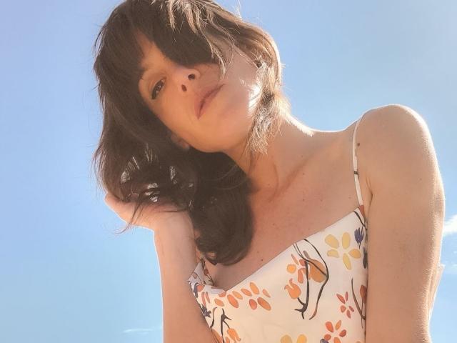 Aiyana with dark wavy hair, wearing a white floral dress, sunlight in her eyes, hand in her hair, blue sky background, looking down with a slight pout, bare shoulders.