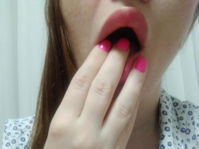 La boca de DashaHart, pintada de rosa, está abierta, mostrando sus dedos, también pintados de rosa, tocando su labio inferior. Lleva una blusa blanca con estampado floral, y se puede ver parte de su cabello castaño..