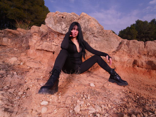 KiraKayleigh, com cabelos longos e pretos, vestindo uma jaqueta preta com zíper e botas, sentada com as pernas abertas em um terreno rochoso, mostrando suas botas pretas e um leve vislumbre de seus seios..