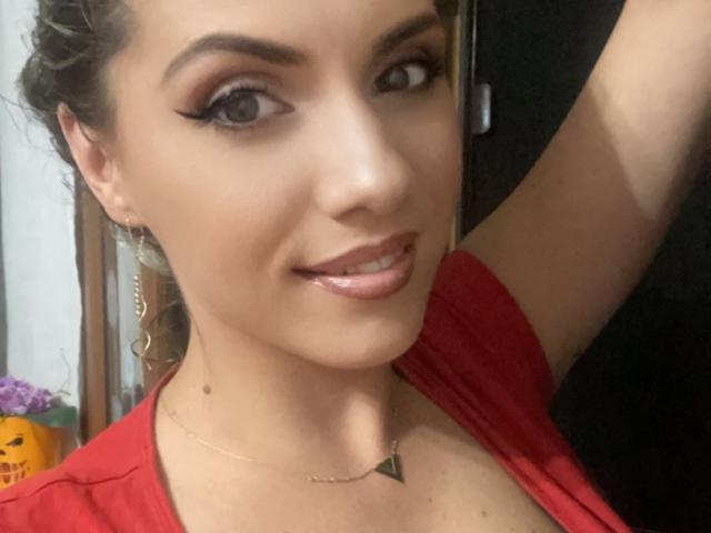 LucilleXSugar has shiny brown eyes, thick black eyeliner, glossy pink lips, and a cute smile. She's wearing a red top and a thin gold necklace with a small triangle pendant.
