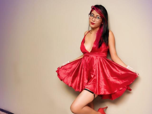 Aby Coleman, vêtue d'une robe rouge, avec un décolleté plongeant, tenant sa jupe, en sous-vêtements noirs, avec des talons rouges, des lunettes, des cheveux roses et noirs, agenouillée, souriante. Les seins sont visibles, les fesses sont légèrement couvertes, le vagin n'est pas visible..