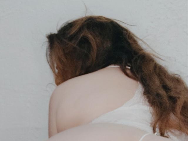 Le dos de Valuna est tourné vers la caméra, révélant ses épaules nues et le haut de son dos. Elle a de longs cheveux bruns et porte un haut blanc, en dentelle, transparent..