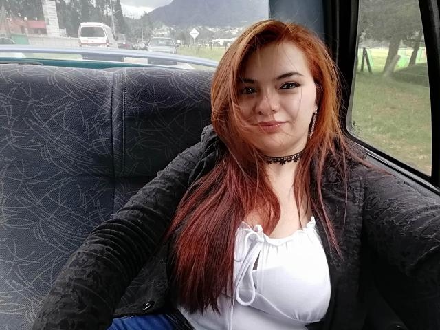KatherinLatin with red hair, black jacket, white top, and choker, sitting in a car, smiling, showing her small tits, inside, with a green landscape outside the window.