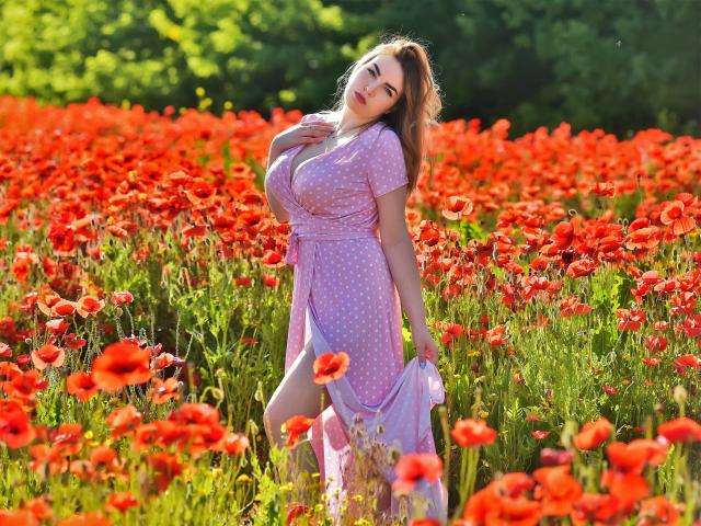 WilsonMelanie em um campo de papoulas vermelhas, vestindo um vestido rosa com bolinhas, segurando um tecido rosa, sorrindo, com os seios levemente visíveis, sob luz solar intensa..