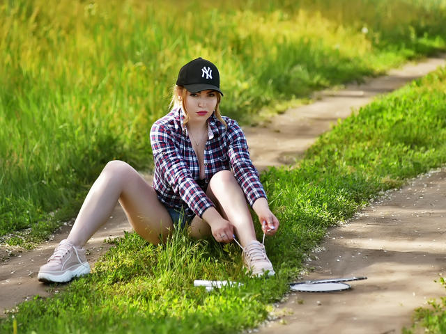 WilsonMelanie est assise sur l'herbe, les jambes écartées, portant une casquette noire "NY", une chemise à carreaux, un short et des baskets, et tenant un stylo, avec un livre sur le chemin..