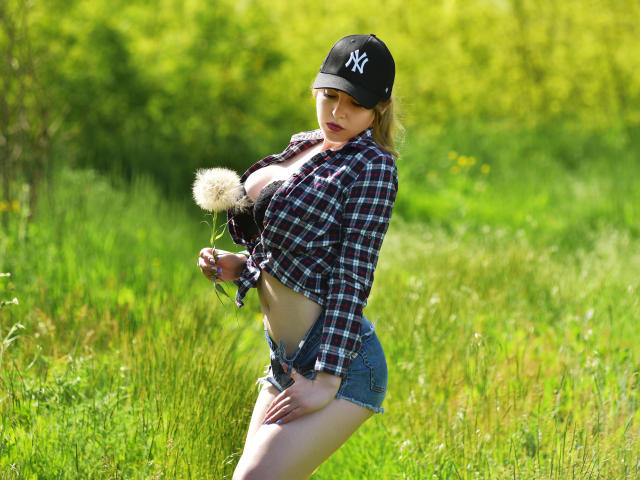 WilsonMelanie, avec de grandes seins, portant une casquette NY, une chemise à carreaux nouée au-dessus, un short en jean, tenant un pissenlit blanc et duveteux, debout dans un champ vert ensoleillé..