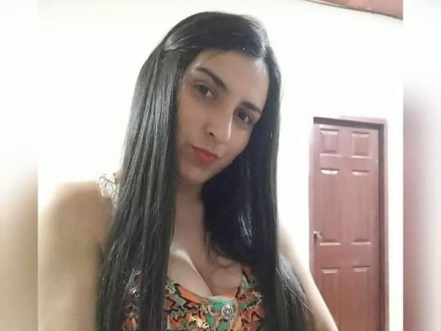 KingaNDher with long black hair, wearing a colorful patterned top, showing small tits, red lips, and a necklace, standing in front of a wooden door.