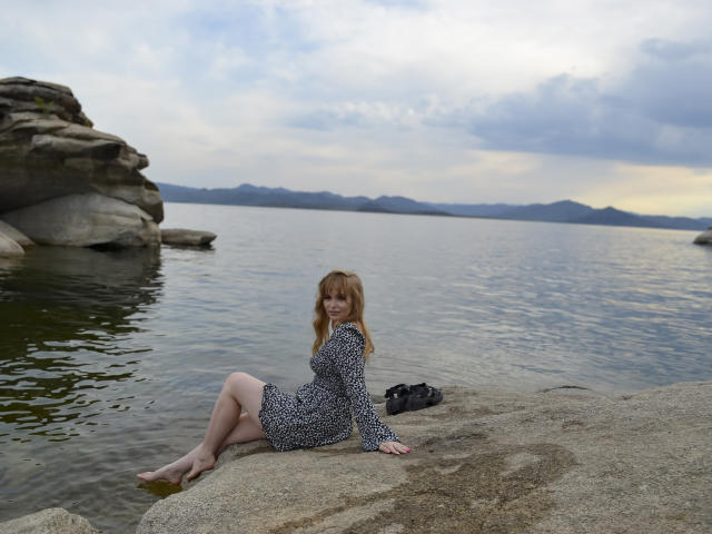 SweetAleksaX, con i capelli rossi, indossa un vestito a stampa leopardo e siede su una roccia vicino all'acqua, con le gambe incrociate, sorridendo. C'è una borsa nera sulla roccia e il cielo è nuvoloso..