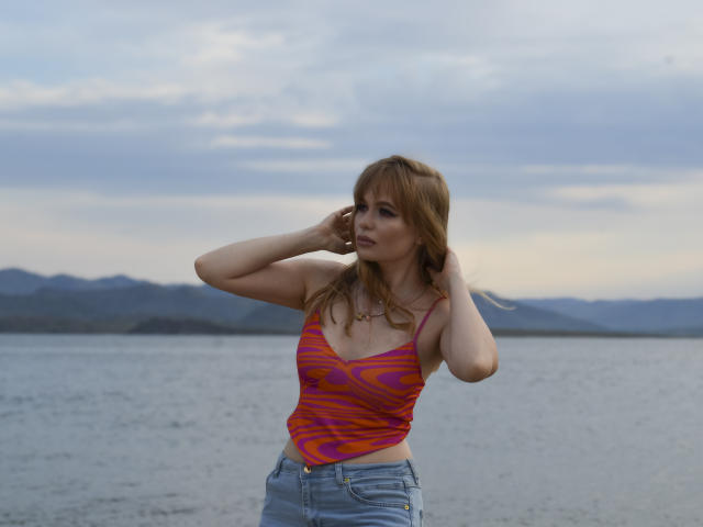 SweetAleksaX has red hair, wearing a pink and orange patterned tank top, and blue jeans, standing by a lake with mountains in the background, hands in her hair.