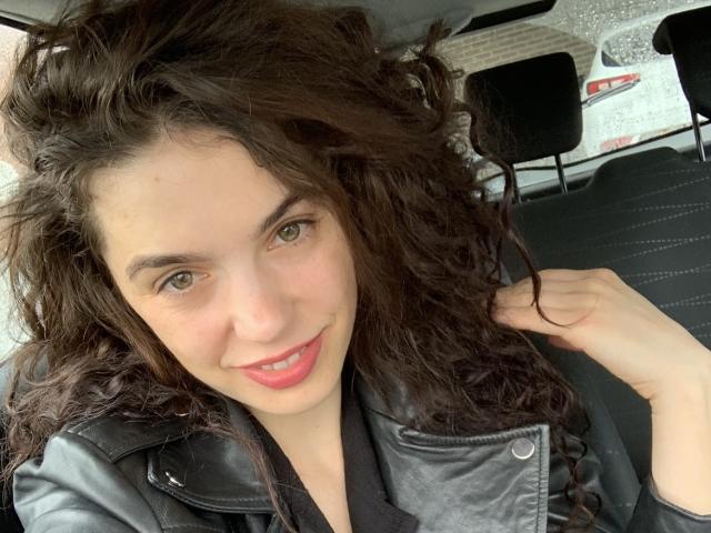 GillKelly with curly brown hair, green eyes, and pink lips, wearing a black leather jacket, smiling in a car, holding her hair with one hand.
