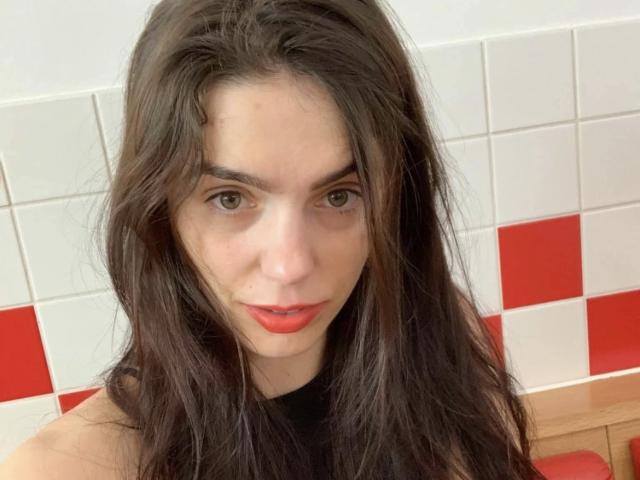 Gill Kelly ha lunghi capelli castani, occhi verdi e rossetto rosso. Sta guardando la fotocamera con un sorriso leggero e sicuro. Le sue natiche non sono visibili..