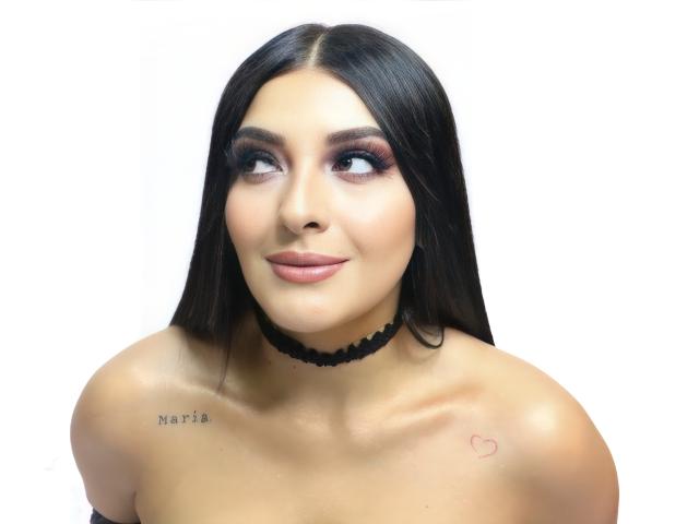 ArianaCortes heeft lang, donker haar, draagt een zwarte choker, kijkt naar de zijkant met roze oogschaduw, en heeft tatoeages op haar rechter schouder en bovenborst. Borsten zichtbaar..