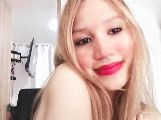 Yvonnex69, met blond haar, een neusring en roze lippenstift, glimlacht naar de camera en laat haar blote schouders en roze lippen zien. Ze heeft lange wimpers en een schattig gezicht..