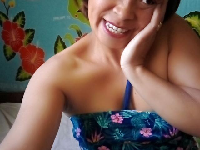 Rahel69, con una sonrisa encantadora, mano en la mejilla, vestido floral azul, mostrando sus senos, piel morena, cabello corto y castaño, y ojos brillantes. Está mirando a la cámara..