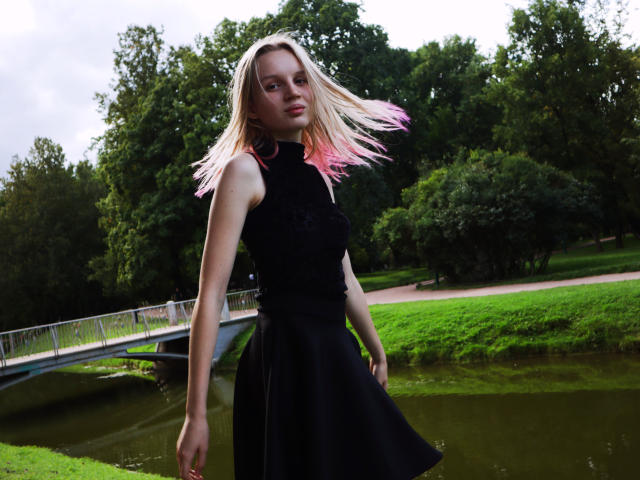 Beatrica Klein, con i capelli biondi con le punte rosa, una maglietta nera senza maniche e una gonna nera, in piedi vicino a un ponte, che guarda verso la fotocamera, con un parco verde sullo sfondo..