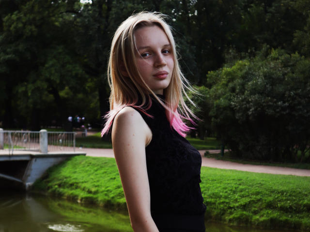 Beatrica Klein tiene el cabello rubio con puntas rosadas, una figura delgada, pechos pequeños, y lleva una blusa negra de encaje sin mangas, de pie junto a un parque con hojas verdes y un canal de agua en el fondo..