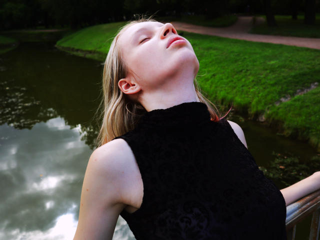 Beatrica Klein, les yeux fermés, la tête penchée en arrière, portant un haut noir en dentelle sans manches, avec des cheveux blonds et une peau pâle, appuyée sur une rambarde près d'un étang réfléchissant..