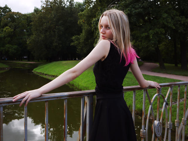 BeatricaKlein with pale skin and pink-tipped blonde hair, wearing a black lace sleeve top and black skirt, leaning on a metal railing, looking back with a slight smirk.