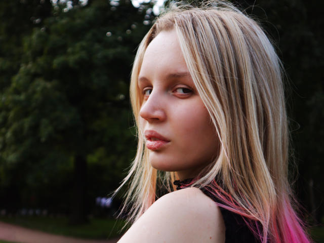 Blond Beatrica Klein, con i capelli dalle punte rosa, labbra piene e un trucco leggero, guarda oltre la spalla. Il suo seno è piccolo e i glutei non sono visibili. Si trova all'aperto..