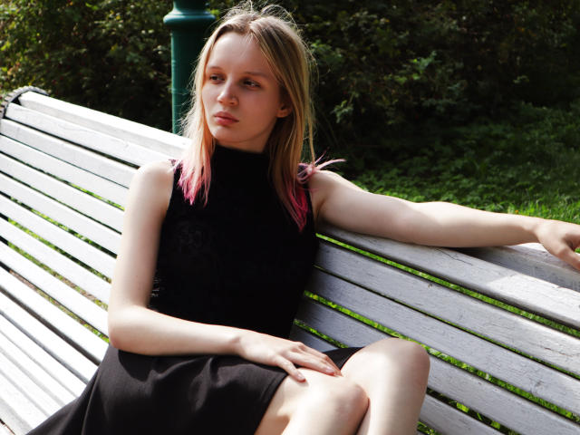 BeatricaKlein with pale skin and pink-tipped blonde hair, wearing a black sleeveless dress, sitting on a wooden bench, arms and legs relaxed, looking slightly bored.