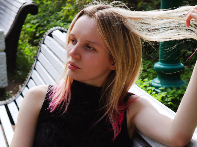 BeatricaKlein with blonde hair, pink tips, holding her hair, wearing a black lace sleeveless top, sitting on a wooden bench, looking off to the side, in a sunny park.