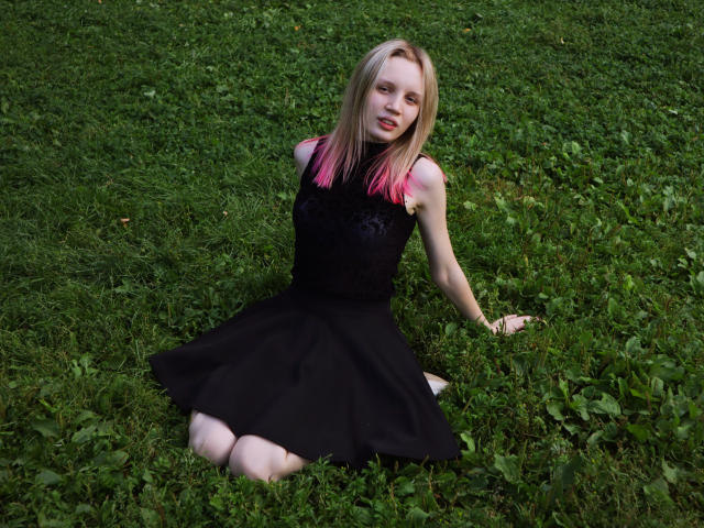 Beatrica Klein, bionda con capelli dalle punte rosa, seduta sull'erba, indossa una canottiera nera in pizzo e una gonna nera, guarda la fotocamera, con le gambe piegate e le mani a terra..