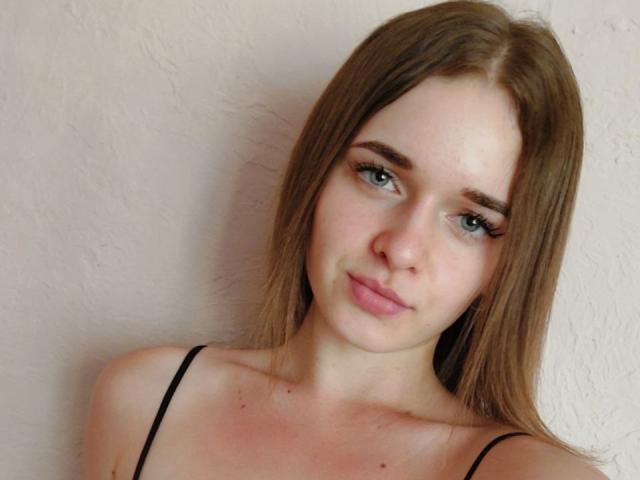 Samanta69Hot has light brown hair, green eyes, and a subtle smile. She's wearing a black spaghetti strap top, showing her small tits and fair skin.
