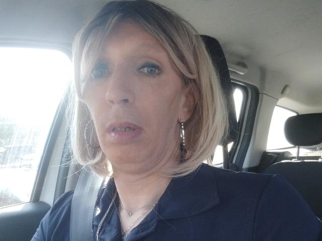 Eine Frau mit blondem Bob, blauen Augen, die goldene Ohrringe trägt und ein dunkelblaues Hemd anhat, sitzt in einem Auto, blickt leicht schmollend in die Kamera und hat den Sicherheitsgurt angelegt..