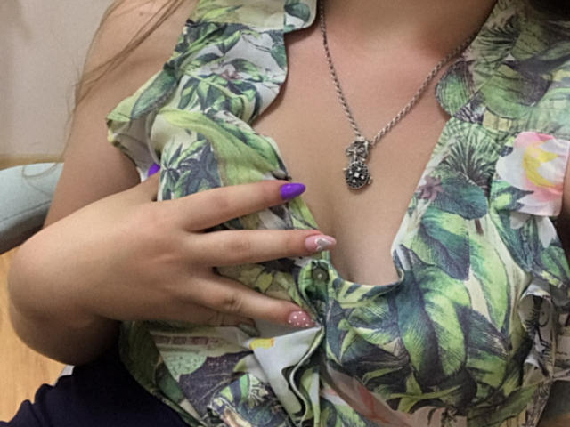La mano de Dooris, con uñas moradas y rosas, toca sus senos, mientras lleva una blusa verde con estampado de hojas, un collar de plata con un colgante negro, y tiene el escote visible..