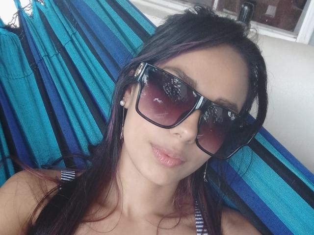 AnaisHilton with dark hair, wearing big black sunglasses, and a striped blue hammock, has pouty pink lips and small earrings, looking cute and relaxed.